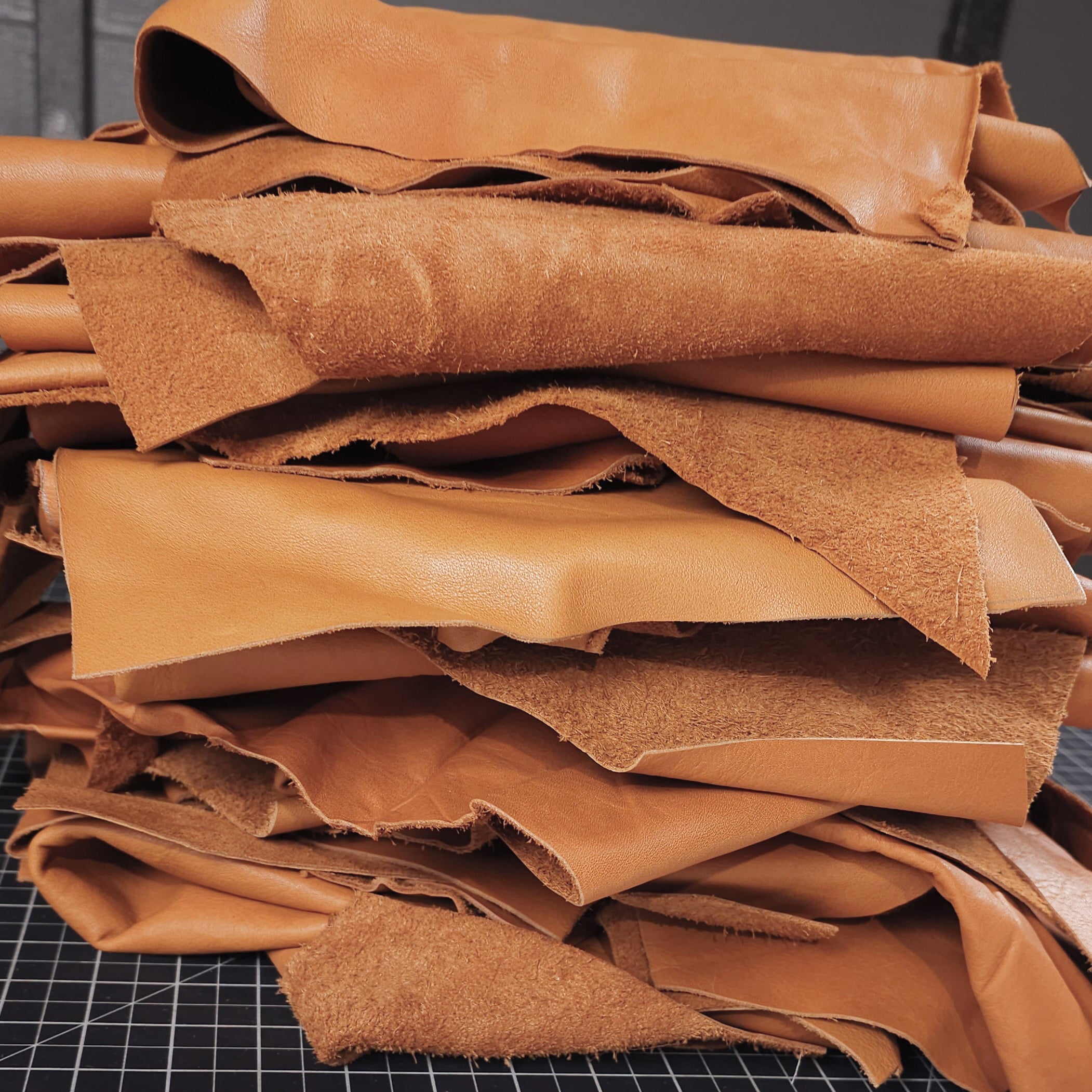 A stack of saddle tan leather remnants with a fine grain texture, displayed on a grid background, indicating they are ready for leathercraft.