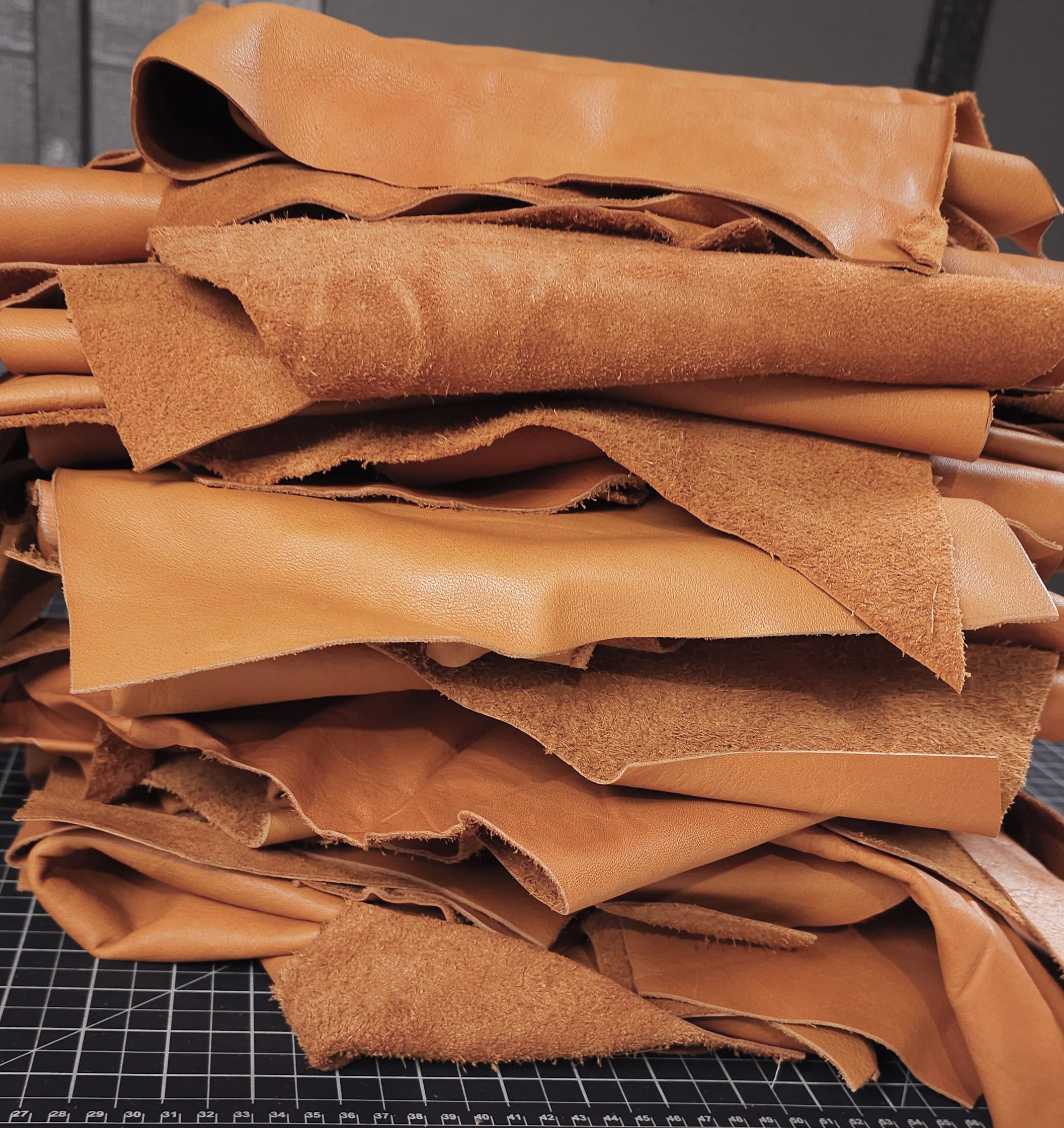 A stack of saddle tan leather remnants with a fine grain texture, displayed on a grid background, indicating they are ready for leathercraft.